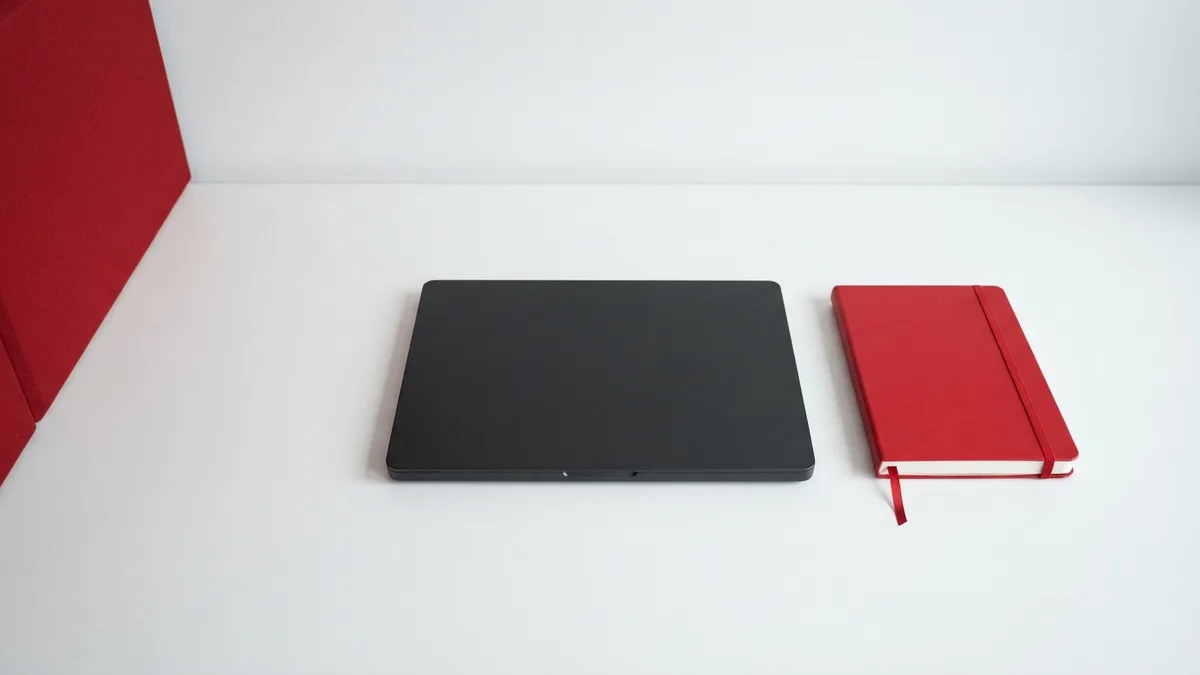 Closed graphite laptop and crimson notebook arranged on a white desk for admin dashboard access