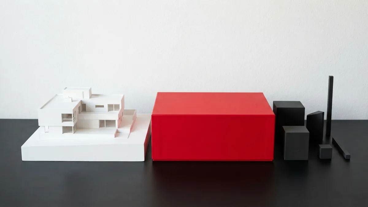Matte red box beside white and graphite geometric forms in a refined minimal scene